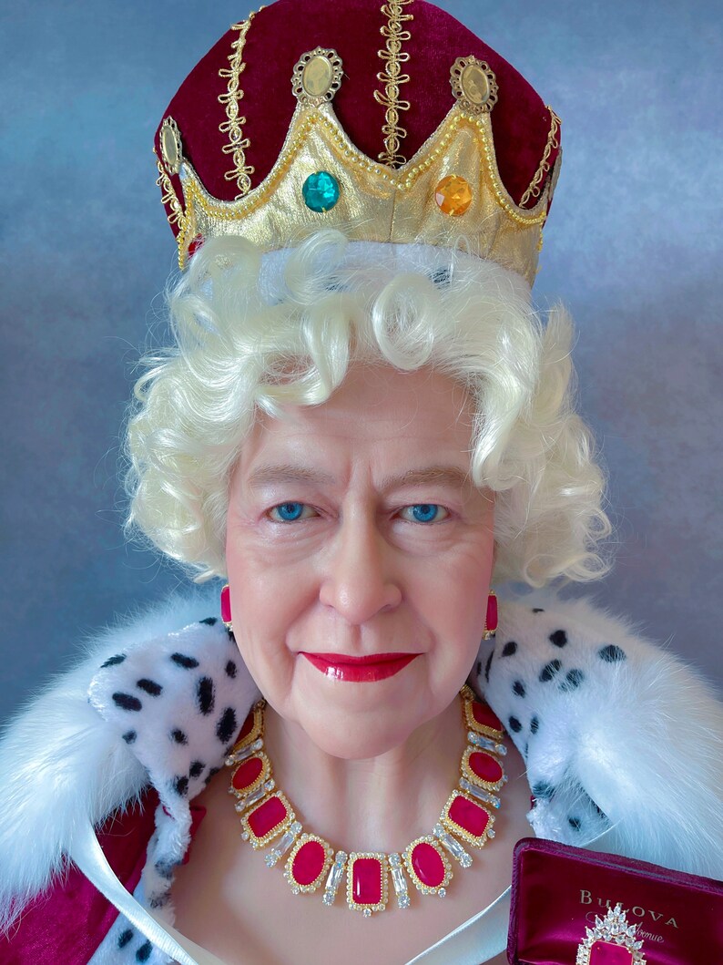 May include: A woman wearing a red velvet crown with gold trim and jewels, a white fur collar, and a red and gold necklace with large pink gemstones. She is wearing a white wig and has a serious expression.