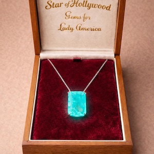 May include: A turquoise gemstone pendant necklace in an open wooden box. The rectangular pendant glows and hangs from a silver chain. The box has a red velvet lining and gold text on the lid that reads "Star of Hollywood Gems for Lady America".