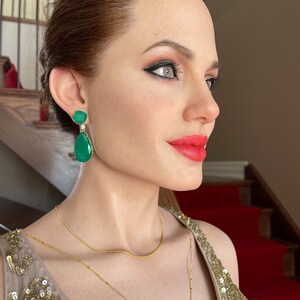 May include: A side profile of a person wearing emerald green drop earrings with a gold necklace and a sequined dress. The person has red lipstick and dark eye makeup. The background includes a staircase with a red carpet.