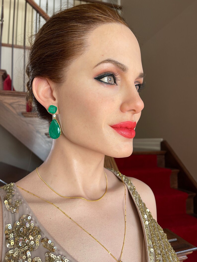 May include: A side profile of a person wearing emerald green drop earrings with a gold necklace and a sequined dress. The person has red lipstick and dark eye makeup. The background includes a staircase with a red carpet.
