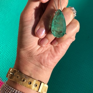 May include: A large, pear-shaped, light green pendant necklace with a silver chain. The pendant is held by a hand, along with a gold mesh bracelet and a diamond-studded bracelet. A diamond ring is also visible.