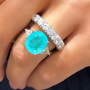 May include: A ring featuring a large, oval, turquoise-coloured gemstone surrounded by smaller, triangular gemstones. The ring is set in silver and is worn on the ring finger of a hand. A second ring with a row of small, round, white gemstones is worn on the same finger.