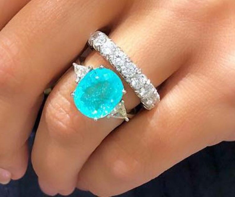 May include: A ring featuring a large, oval, turquoise-coloured gemstone surrounded by smaller, triangular gemstones. The ring is set in silver and is worn on the ring finger of a hand. A second ring with a row of small, round, white gemstones is worn on the same finger.