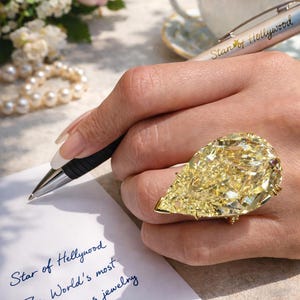 Puede incluir: Un gran anillo de diamantes amarillos en forma de pera. El anillo está en una mano que sostiene un bolígrafo, escribiendo en un trozo de papel. El bolígrafo es plateado y negro. El papel tiene la inscripción "Star of Hollywood".