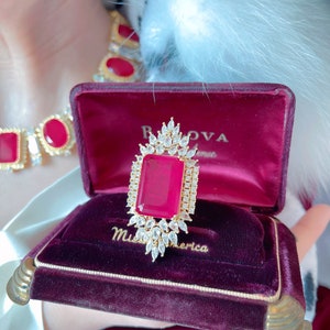 May include: A red velvet ring box with gold trim, featuring a large red gemstone ring with a diamond halo setting. The box is inscribed with "Miss America" and "B.O.V.A.  Avenue".