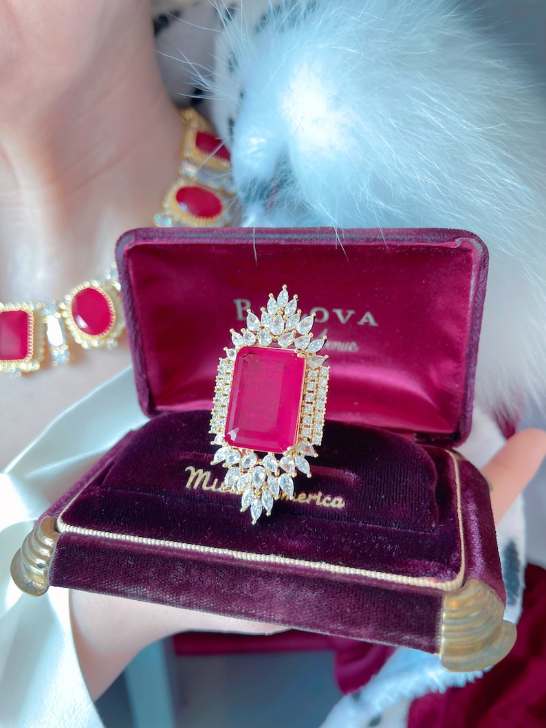 May include: A red velvet ring box with gold trim, featuring a large red gemstone ring with a diamond halo setting. The box is inscribed with "Miss America" and "B.O.V.A.  Avenue".