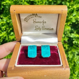 May include: A wooden box with a red velvet interior, containing a pair of turquoise blue earrings. The box has the text "Columbia Diamond Rings Gems for Lady America" on the lid.