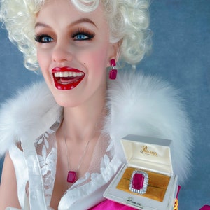 May include: A woman with blonde hair is wearing a white top with lace detailing and a pink glove. She is holding a small white box with a red jewel ring inside. The box has the text "Princess" on it.