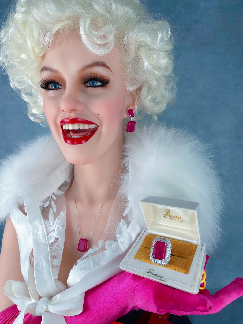 May include: A woman with blonde hair is wearing a white top with lace detailing and a pink glove. She is holding a small white box with a red jewel ring inside. The box has the text "Princess" on it.