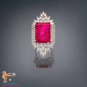 May include: A silver ring featuring a large, rectangular, pink gemstone surrounded by smaller, clear gemstones. The ring is set on a silver band.