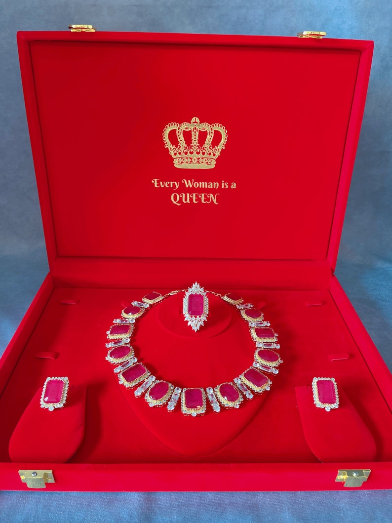May include: A red velvet jewelry box with a gold crown and the text "Every Woman is a Queen". The box contains a ruby and diamond necklace, a pair of earrings, and a pendant.