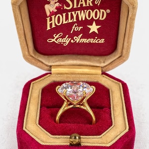May include: A gold ring with a large, clear gemstone, displayed in a red velvet box. The box is inscribed with "Star of Hollywood for Lady America" in gold lettering. The box has gold trim and a small clasp.