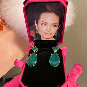 May include: A pink velvet jewelry box with a black interior. The box is open and contains a pair of green teardrop-shaped earrings with gold accents. A photo of a woman is visible inside the box.
