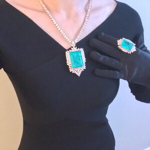 Op de afbeelding: Een zwarte jurk met een grote smaragdgroene edelsteen hangerketting en een ring met een vergelijkbare smaragdgroene edelsteen. De ring zit op een zwarte satijnen handschoen.