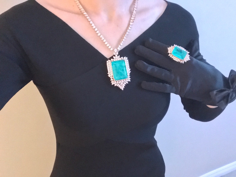 Op de afbeelding: Een zwarte jurk met een grote smaragdgroene edelsteen hangerketting en een ring met een vergelijkbare smaragdgroene edelsteen. De ring zit op een zwarte satijnen handschoen.