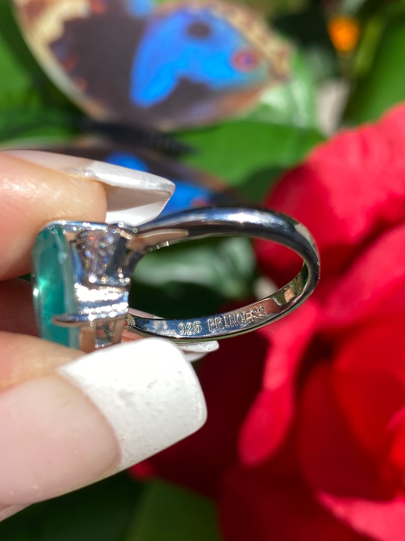 May include: A silver ring featuring a large, oval-shaped green gemstone and a smaller, round clear gemstone. The ring is marked "925 Princess" on the inside band.