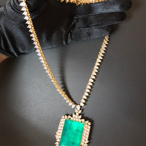 Op de afbeelding: Een gouden ketting met een grote hanger van smaragdgroene edelsteen, omringd door kleinere heldere edelstenen.