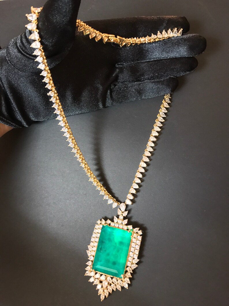 Op de afbeelding: Een gouden ketting met een grote hanger van smaragdgroene edelsteen, omringd door kleinere heldere edelstenen.