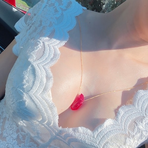 May include: A gold chain necklace with a pink heart-shaped pendant. The necklace is worn by a person wearing a white lace top.