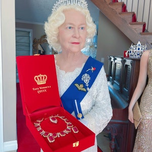 Puede incluir: Una mujer con un vestido blanco y una corona sostiene una caja de terciopelo rojo con una corona dorada y las palabras "Every Woman is a Queen" en la tapa. La caja está abierta y contiene un collar de rubíes y diamantes.