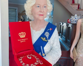 May include: A woman wearing a white dress and a crown holds a red velvet box with a gold crown and the words "Every Woman is a Queen" on the lid. The box is open and contains a ruby and diamond necklace.