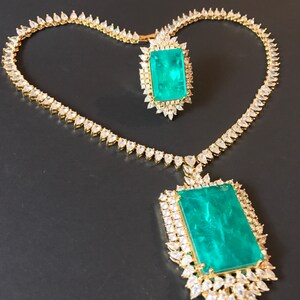 Op de afbeelding: Een gouden ketting- en ringset met een grote smaragdgroene edelsteen omringd door kleinere heldere edelstenen.
