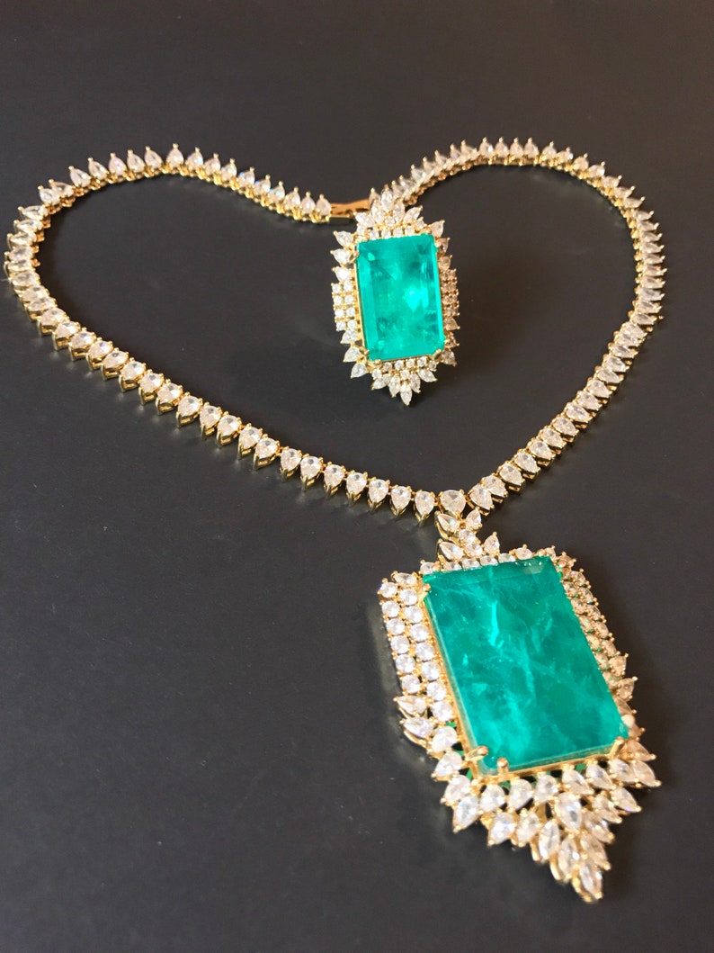 Op de afbeelding: Een gouden ketting- en ringset met een grote smaragdgroene edelsteen omringd door kleinere heldere edelstenen.