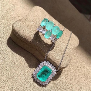 May include: A silver necklace with a large emerald green gemstone pendant surrounded by smaller clear gemstones. The pendant is in a rectangular shape. The necklace is on a light brown surface. There is also a silver ring with multiple emerald green gemstones set in silver.