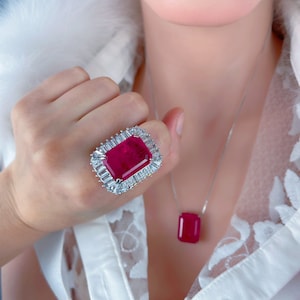 May include: A silver ring with a large, square, red gemstone surrounded by smaller, clear gemstones. The ring is worn on the index finger of a hand. A silver necklace with a matching red gemstone pendant is also visible.
