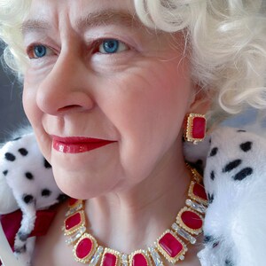 May include: A close-up of a woman wearing a white fur coat and a gold necklace with large red gemstones. She is also wearing a pair of red gemstone earrings.