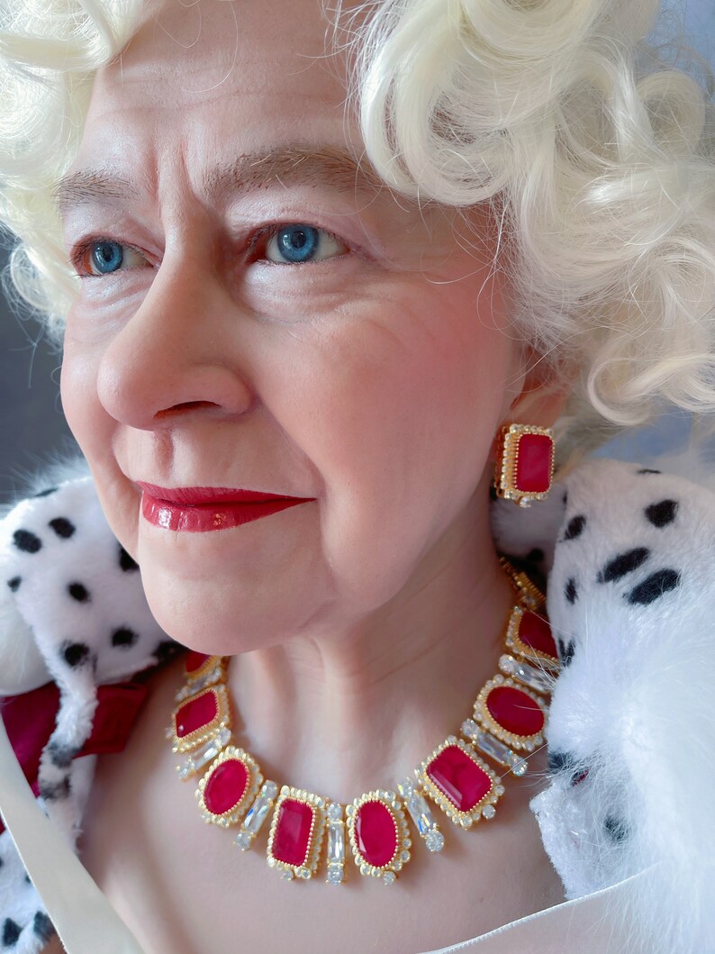 May include: A close-up of a woman wearing a white fur coat and a gold necklace with large red gemstones. She is also wearing a pair of red gemstone earrings.