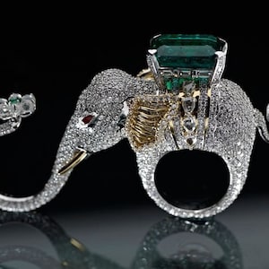 May include: A silver ring shaped like an elephant with a large green emerald gemstone on its back. The elephant is covered in small diamonds.