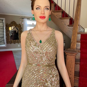 May include: A wax figure of a woman wearing a gold sequined gown with a V-neckline. She is adorned with emerald green earrings and a matching pendant necklace. Her red lips and light skin contrast with the gold and green accents. The figure is standing on a red carpet.