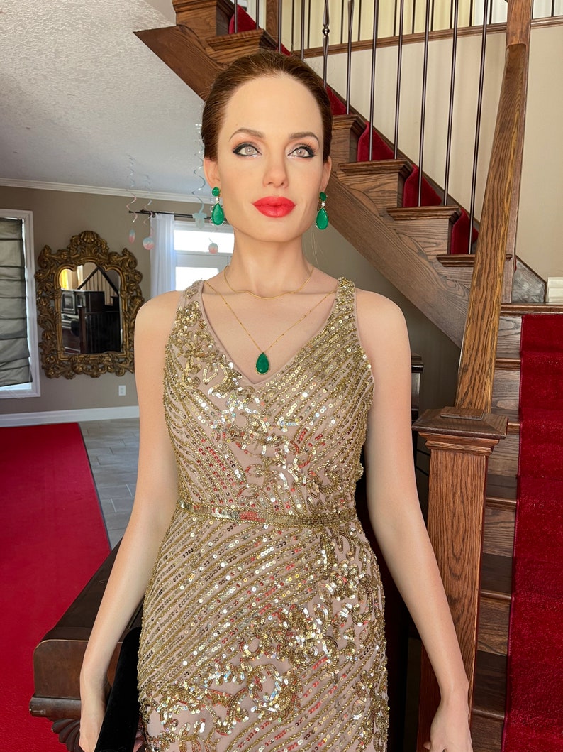 May include: A wax figure of a woman wearing a gold sequined gown with a V-neckline. She is adorned with emerald green earrings and a matching pendant necklace. Her red lips and light skin contrast with the gold and green accents. The figure is standing on a red carpet.