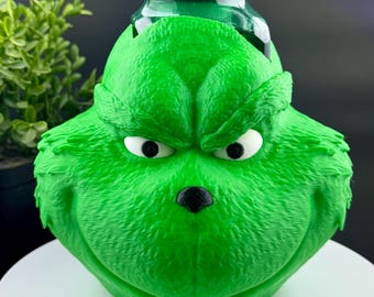 Könnte beinhalten: Ein grüner Flaschenaufsatz im Grinch-Design. Der Aufsatz zeigt das Gesicht des Grinch mit weißen Augen, einer schwarzen Nase und einem wütenden Gesichtsausdruck. Der grüne Verschluss der Flasche ist sichtbar.