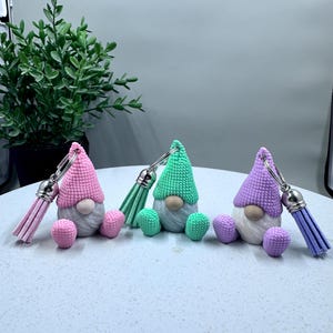May include: Three gnome keychains with different colored hats and tassels. The gnomes are wearing pink, green, and purple hats. The tassels are pink, green, and purple.