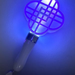 May include: A white and silver handheld device with a four-leaf clover-shaped head, illuminated with blue light. The handle is white and has a wrist strap. The head has a grid pattern.