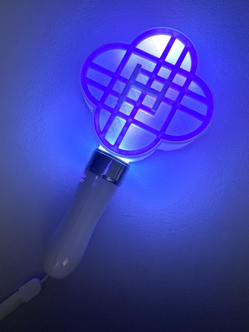 May include: A white and silver handheld device with a four-leaf clover-shaped head, illuminated with blue light. The handle is white and has a wrist strap. The head has a grid pattern.
