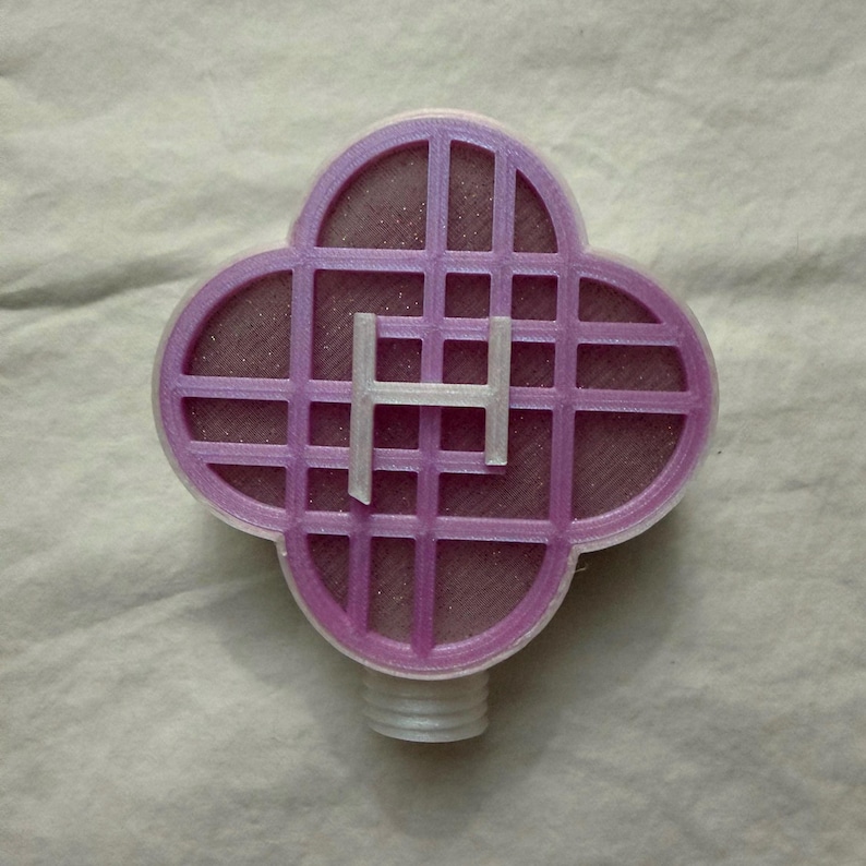 May include: A lavender-coloured, four-leaf clover-shaped object with a grid pattern and a white letter "H" in the centre. The object has a threaded base. The item is likely a decorative or functional piece.