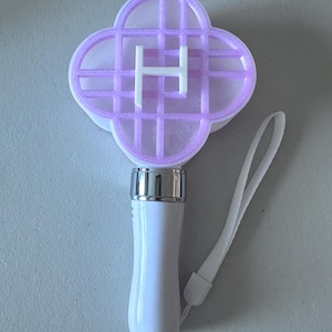 May include: A white and silver light stick with a four-leaf clover-shaped head. The head is lavender with a white "H" in the center. A white wrist strap is attached to the base of the handle.