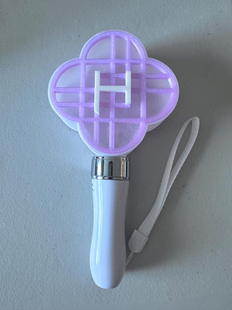 May include: A white light stick with a silver base and a four-leaf clover-shaped head. The head is lavender with a white "H". A white wrist strap is attached to the base of the handle.