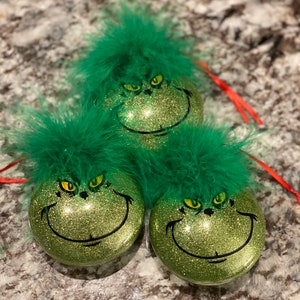 May include: Three green Grinch Christmas ornaments with glitter and a fuzzy green feather trim. Each ornament features a Grinch face with yellow eyes and a black smile.