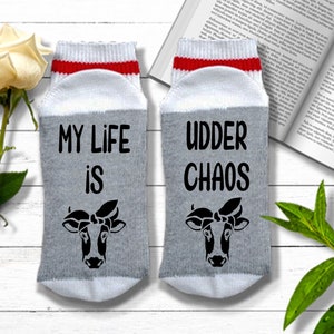 K&ouml;nnte beinhalten: Ein Paar graue und wei&szlig;e Socken mit rotem Besatz. Eine Socke sagt "My life is" mit einer Kuh-Illustration. Die andere Socke sagt "Udder Chaos" mit einer Kuh-Illustration.