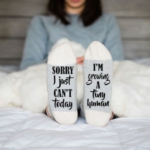 May include: A pair of white socks with gray heels and toes. One sock reads "Sorry I just can't today" and the other reads "I'm growing a tiny human".