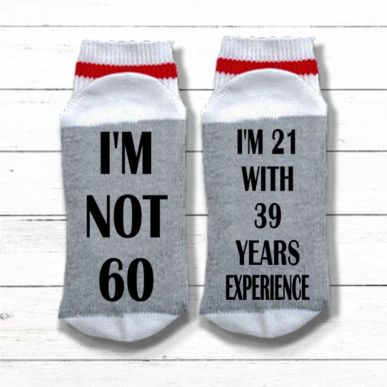 May include: A pair of gray and white socks with red stripes. One sock says "I'm not 60" and the other sock says "I'm 21 with 39 years experience".