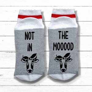 K&ouml;nnte beinhalten: Ein Paar graue und wei&szlig;e Socken mit roten Streifen. Eine Socke sagt "NOT in" und die andere sagt "THE MOOOOD". Beide Socken haben ein schwarz-wei&szlig;es Kuhgesicht mit einer Schleife.