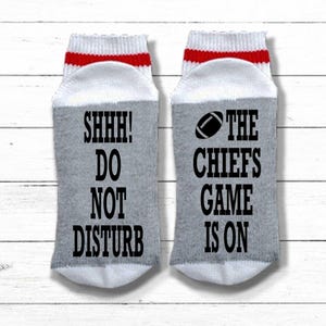 Peut inclure: Une paire de chaussettes grises avec des hauts blancs et des rayures rouges. Une chaussette porte l'inscription "SHHH! DO NOT DISTURB" en noir. L'autre chaussette porte un dessin de ballon de football et l'inscription "THE CHIEFS GAME IS ON".
