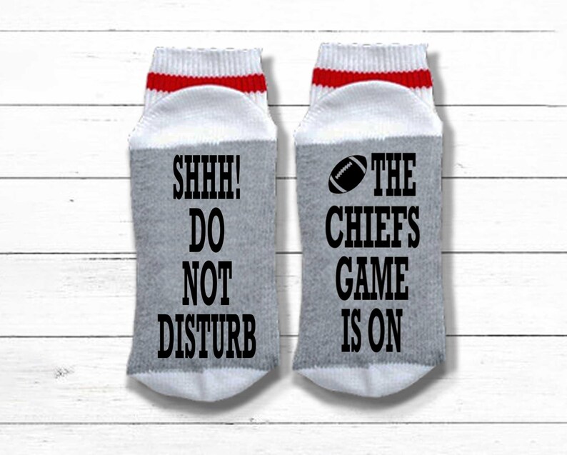Peut inclure: Une paire de chaussettes grises avec des hauts blancs et des rayures rouges. Une chaussette porte l'inscription "SHHH! DO NOT DISTURB" en noir. L'autre chaussette porte un dessin de ballon de football et l'inscription "THE CHIEFS GAME IS ON".