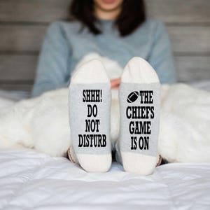 Peut inclure: Paire de chaussettes grises et blanches avec l'inscription "SHHH! DO NOT DISTURB" et "THE CHIEFS GAME IS ON" en noir. Les chaussettes comportent un motif de ballon de football. Les chaussettes sont sur un lit blanc.