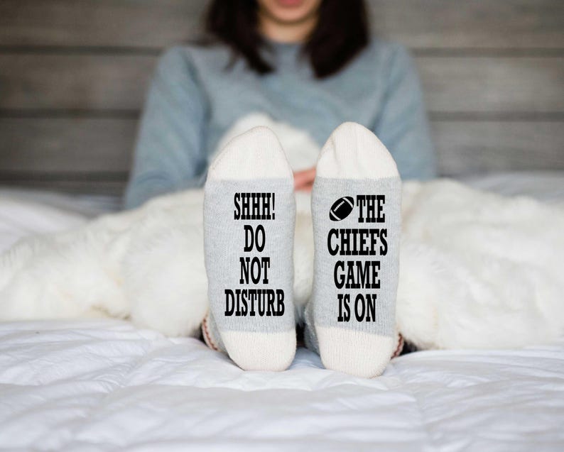 Peut inclure: Paire de chaussettes grises et blanches avec l'inscription "SHHH! DO NOT DISTURB" et "THE CHIEFS GAME IS ON" en noir. Les chaussettes comportent un motif de ballon de football. Les chaussettes sont sur un lit blanc.